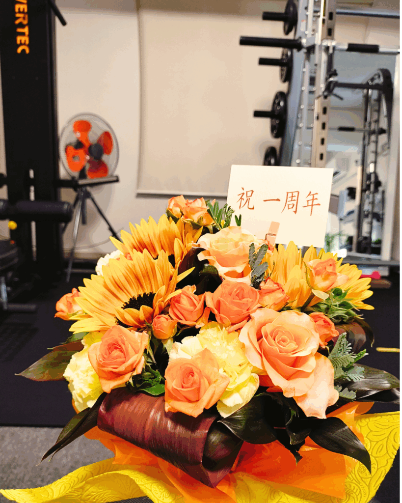 店内のトレーニングエリアに飾られた、ひまわりとオレンジ色の花々の「祝一周年」花束。